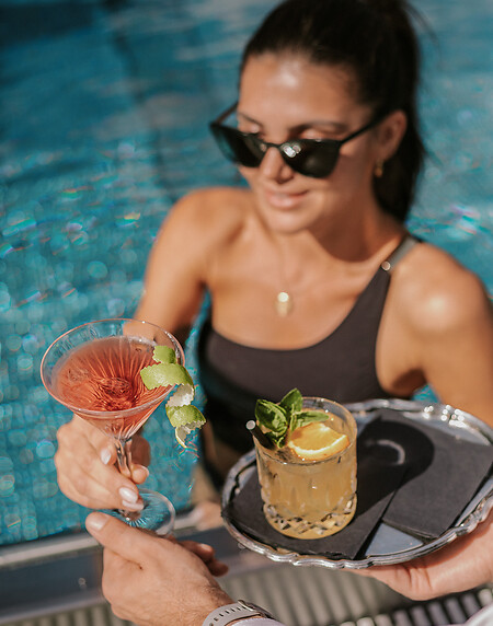 Frau mit Sonnenbrille im Pool bekommt Getränke, Cocktails auf silbernem Tablett geliefert