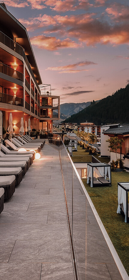 Hotelterrasse mit Sonnenliegen bei Sonnenuntergang – darunter beleuchtete Gebäude im Tal und umliegende Berge.