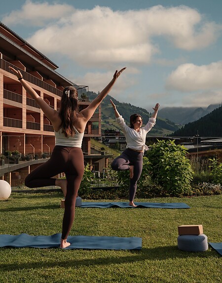 Zwei Frauen stehen auf Yogamatten im Freien und machen eine Baum-Pose mit erhobenen Armen vor einem Hotelgebäude und Bergen im Hintergrund.