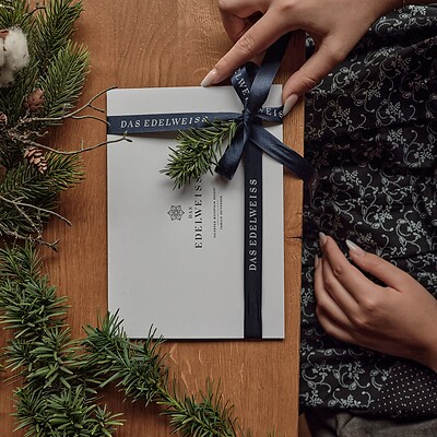 Hände binden ein dunkelblaues Band mit dem Schriftzug 'DAS EDELWEISS' um eine weiße Karte auf einem Holztisch neben Tannenzweigen.