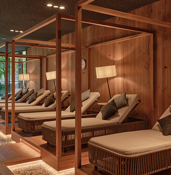Ruhiger Wellnessbereich mit Holzliegen, Vorhängen und warmem Lichtdesign, ausgelegt mit textilen Auflagen.