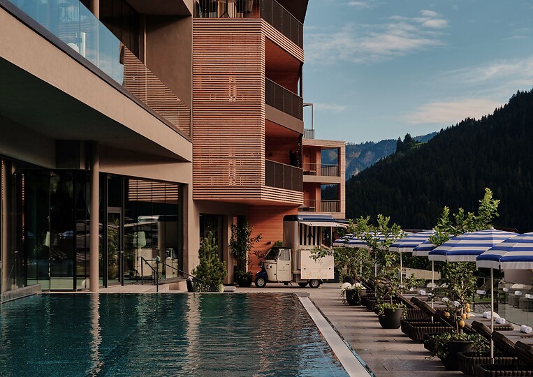 Ein großer Außenpool verläuft entlang der modernen Hotelfassade - rechts stehen Sonnenliegen und Schirme, dahinter Berge und bewaldete Hänge unter blauem Himmel.