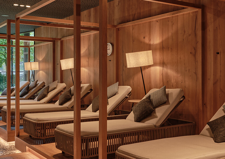 Ruhiger Wellnessbereich mit Holzliegen, Vorhängen und warmem Lichtdesign, ausgelegt mit textilen Auflagen.
