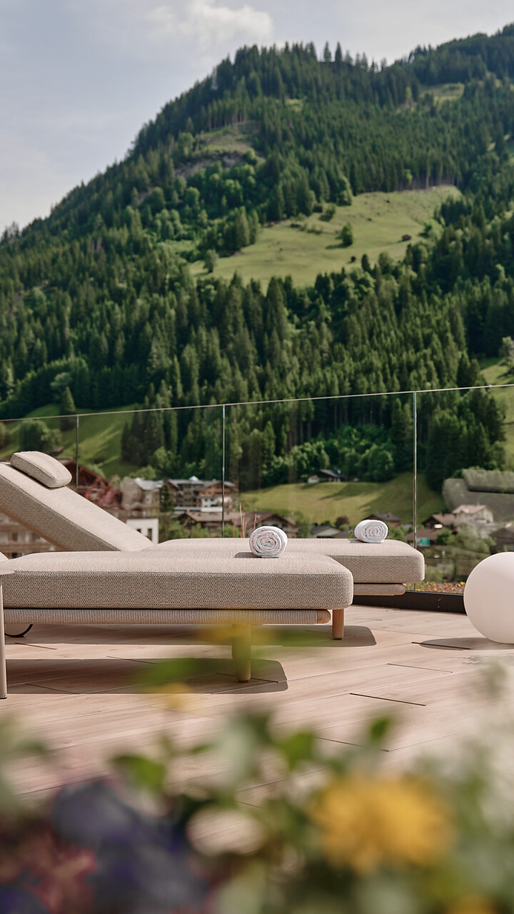 Zwei Sonnenliegen und runde Outdoor-Leuchten stehen auf einer großen Terrasse, dahinter ein grüner Hang mit dichtem Wald und klarer Bergkulisse.