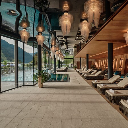 Langer Ruheraum mit Liegen, warmem Holzinterieur und dekorativen Hängelampen – links eine Glasfront mit Blick auf einen Pool und Berge.