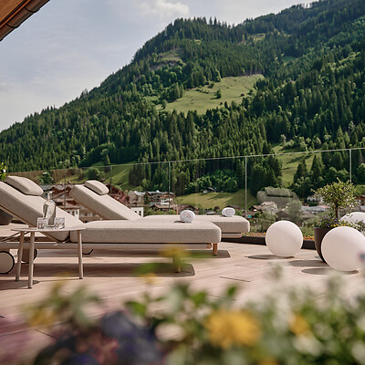 Zwei Sonnenliegen und runde Outdoor-Leuchten stehen auf einer großen Terrasse, dahinter ein grüner Hang mit dichtem Wald und klarer Bergkulisse.