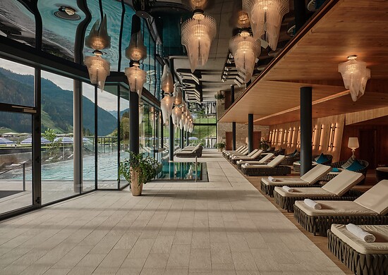Langer Ruheraum mit Liegen, warmem Holzinterieur und dekorativen Hängelampen – links eine Glasfront mit Blick auf einen Pool und Berge.