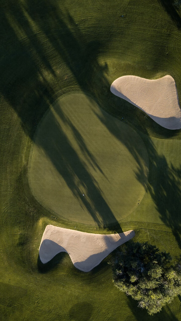 Luftaufnahme eines Golfplatzes mit zwei Sandbunkern und mehreren Bäumen, die lange Schatten werfen.