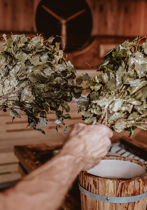 Zwei Hände halten jeweils einen Birkenreisigbesen in einer Holzsauna.