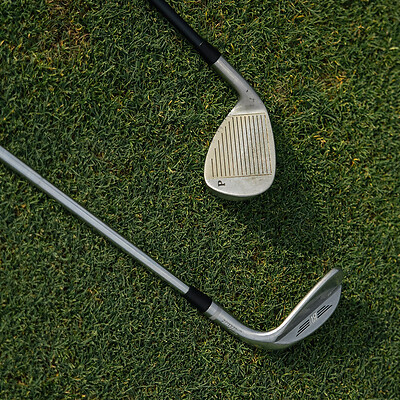 Zwei Golfschläger liegen auf grünem Rasen, einer mit sichtbaren Rillen auf der Schlagfläche.