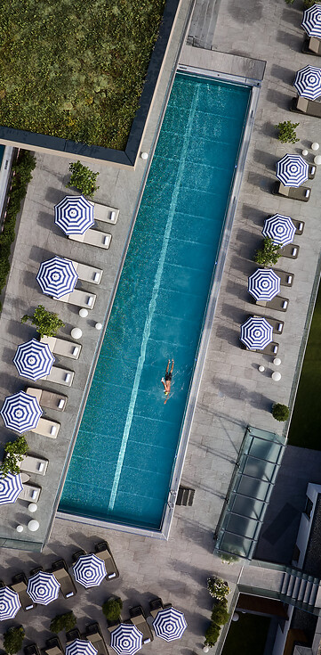 Luftaufnahme eines rechteckigen Außenpools mit mehreren Liegestühlen und Sonnenschirmen sowie einer Person im Wasser neben einem weiteren kleineren Pool und umgeben von Rasenflächen mit Sitzgelegenheiten.