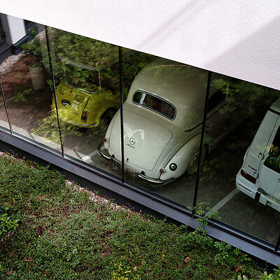 Drei geparkte Autos hinter einer Glasfassade, darunter ein weißer Oldtimer mit Mercedes-Stern, ein gelbes kleines Auto und ein weißer Geländewagen.