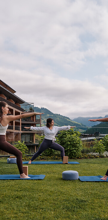 Drei Frauen auf Yogamatten führen im Freien vor einem Hotelgebäude eine Yogapose aus.