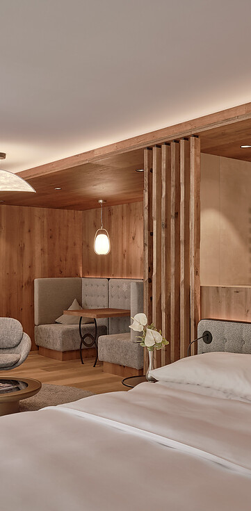 Hotelzimmer mit Doppelbett, zwei grauen Sesseln vor runden Tischen, Holzwänden und integrierter Sitzecke mit Polsterbank.