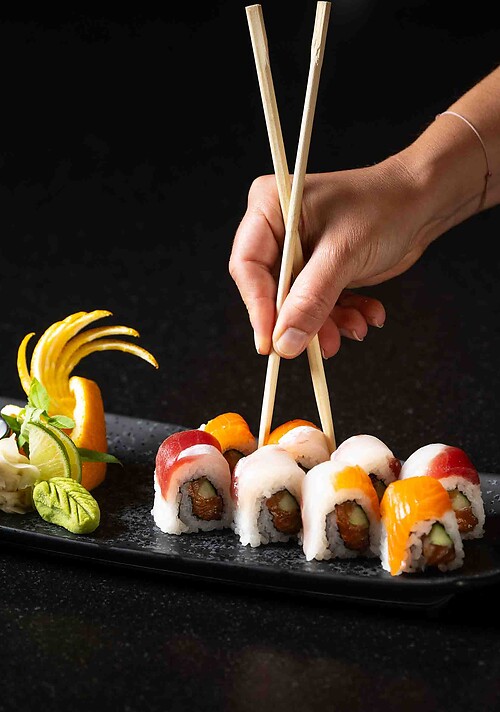 Ein rechteckiger dunkler Teller mit arrangiertem Sushi und einer Garnitur aus Zitrusfrucht und Kräutern auf schwarzem Untergrund - während eine Hand die Stäbchen hält danach greift.