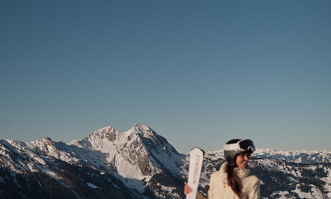 Skifahrerin mit Skiern in der Hand steht auf schneebedecktem Berg mit Alpenpanorama im Hintergrund.