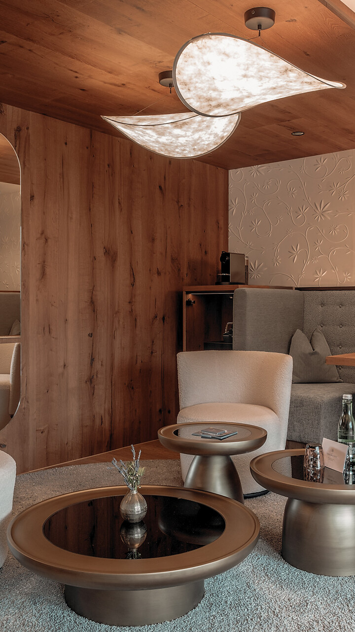 Wohnzimmer mit zwei runden Sesseln, drei runden Tischen auf einem runden Teppich, einer grauen Eckbank mit Tisch und einer Holzvertäfelung mit Spiegel.